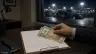 يد تضع أوراق دولار من فئة 100 وأوراق ريال سعودي على لوح مشبك فوق فاتورة شحن بلا كتابة مقروءة في مكتب صغير قرب ميناء جدة ليلًا والحاويات تظهر من النافذة