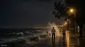 أمواج تتكسر على حاجز كورنيش السويس ليلًا تحت أعمدة الإنارة مع رصيف مبلل ومارة يظهرون من الخلف في ملابس خفيفة ورياح تحرك الأشجار